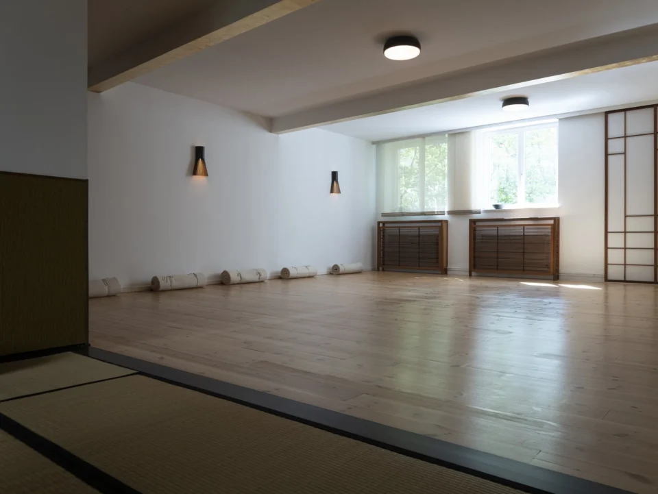 Ein sanft beleuchteter Yogaraum mit Holzboden und aufgerollten weißen Matten an der Wand.